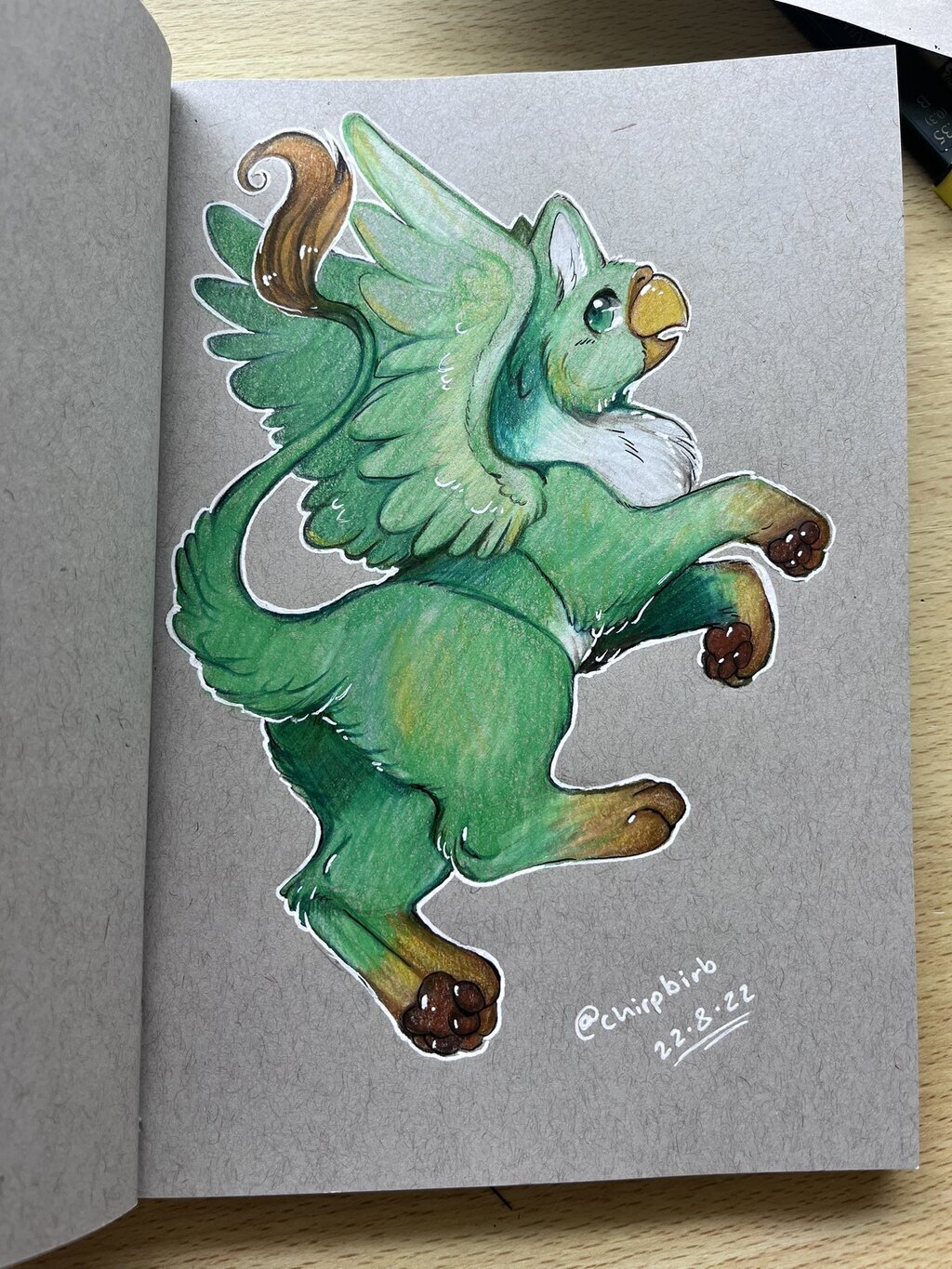 A green gryphon with a white belly and brown paws poses while looking at the camera. Artwork by ApesLion. Colored pencil on textured paper.